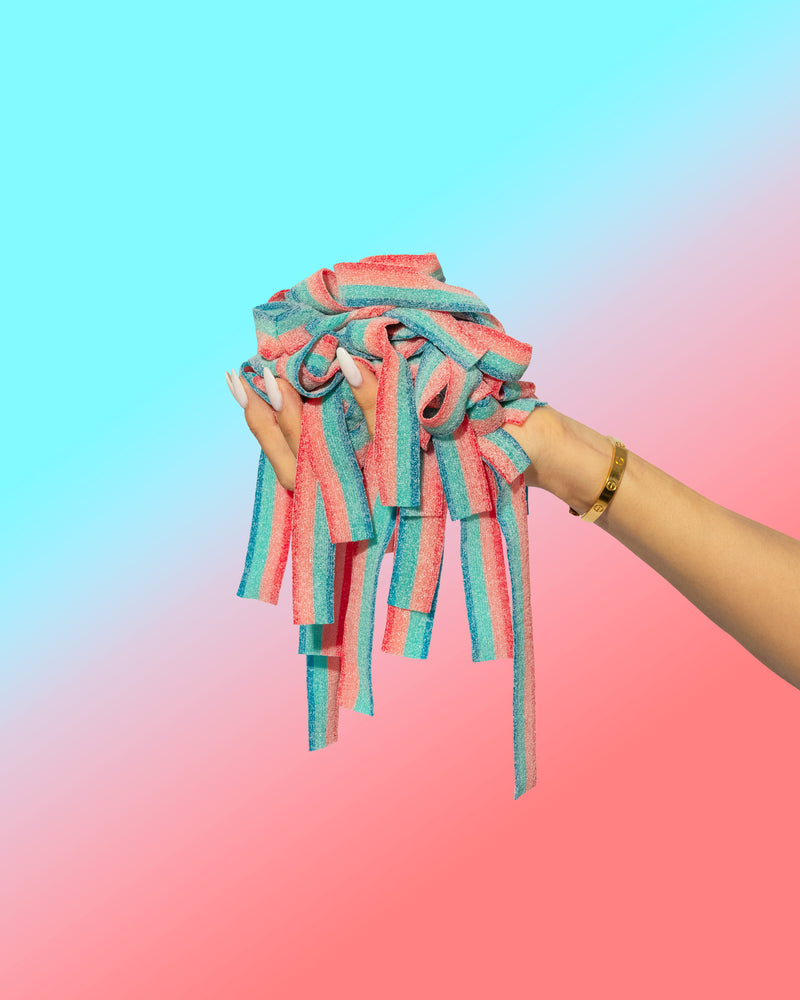 A hand grips colorful pink and blue gummy candies from a Doubleberry-Six Pack by SourStrips against a gradient backdrop of light blue to pink. The person, wearing a gold bracelet, enjoys the intensely sour taste.