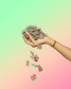 A hand with long nails and a gold bracelet drops colorful Sour Strips Bites-Four Pack red and green candies from Sour Strips against a pastel gradient background.