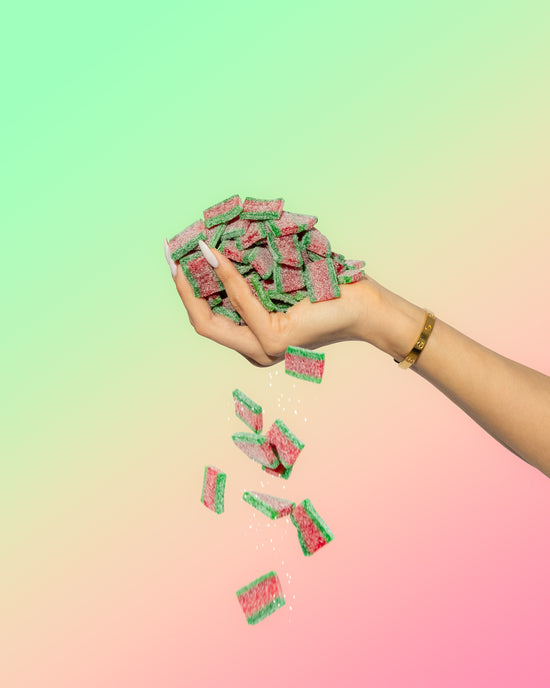 A hand with long nails and a gold bracelet drops several green and pink SourStrips from the Summer Sampler Pack-five pack against a pastel gradient background, showcasing the tangy summer flavors of this SourStrips treat.