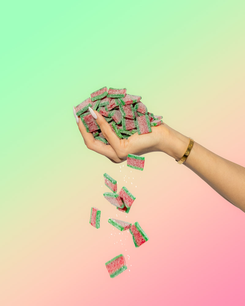 A hand with long nails and a gold bracelet drops several green and pink SourStrips from the Summer Sampler Pack-five pack against a pastel gradient background, showcasing the tangy summer flavors of this SourStrips treat.