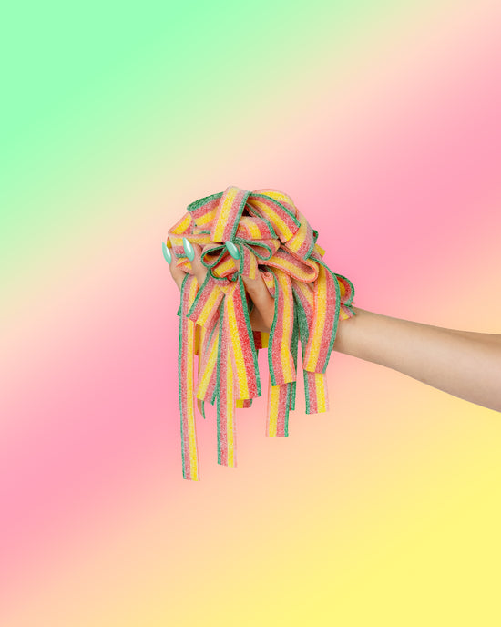 A hand holds a bunch of colorful SourStrips from the Summer Sampler Pack-five pack against a pastel green, pink, and yellow background—perfect for a fun picnic snack.