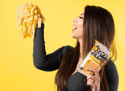 A woman with long dark hair, wearing a black top, is holding a bunch of yellow sour candy strips in one hand and a package labeled "Sour Strips" in the other. She is tilting her head back with her mouth open as if about to eat the candy. The background is bright yellow.