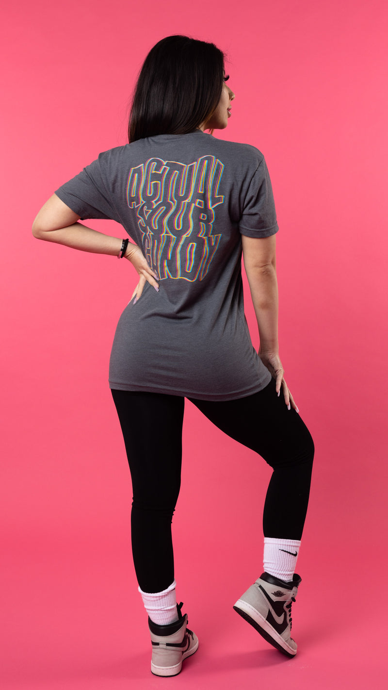 A person with long dark hair stands facing away on a pink background. They wear a grey graphic T-shirt called the "Trippy Tee" by SourStrips, black leggings, white socks, and grey sneakers. Their left hand is on their hip and their right hand rests on their upper right thigh.