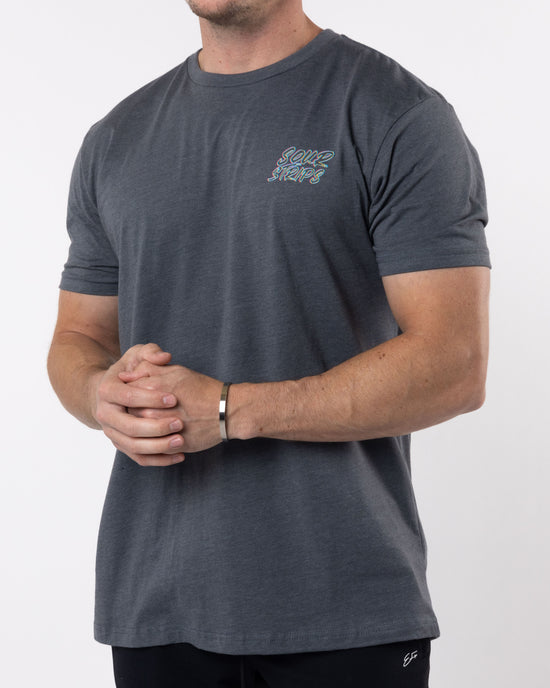 A person wearing a super soft cotton gray "Trippy Tee" by SourStrips, featuring the words "Some Stars" in colorful text on the chest. They have their hands clasped together and are also sporting a silver bracelet. The background is plain white.