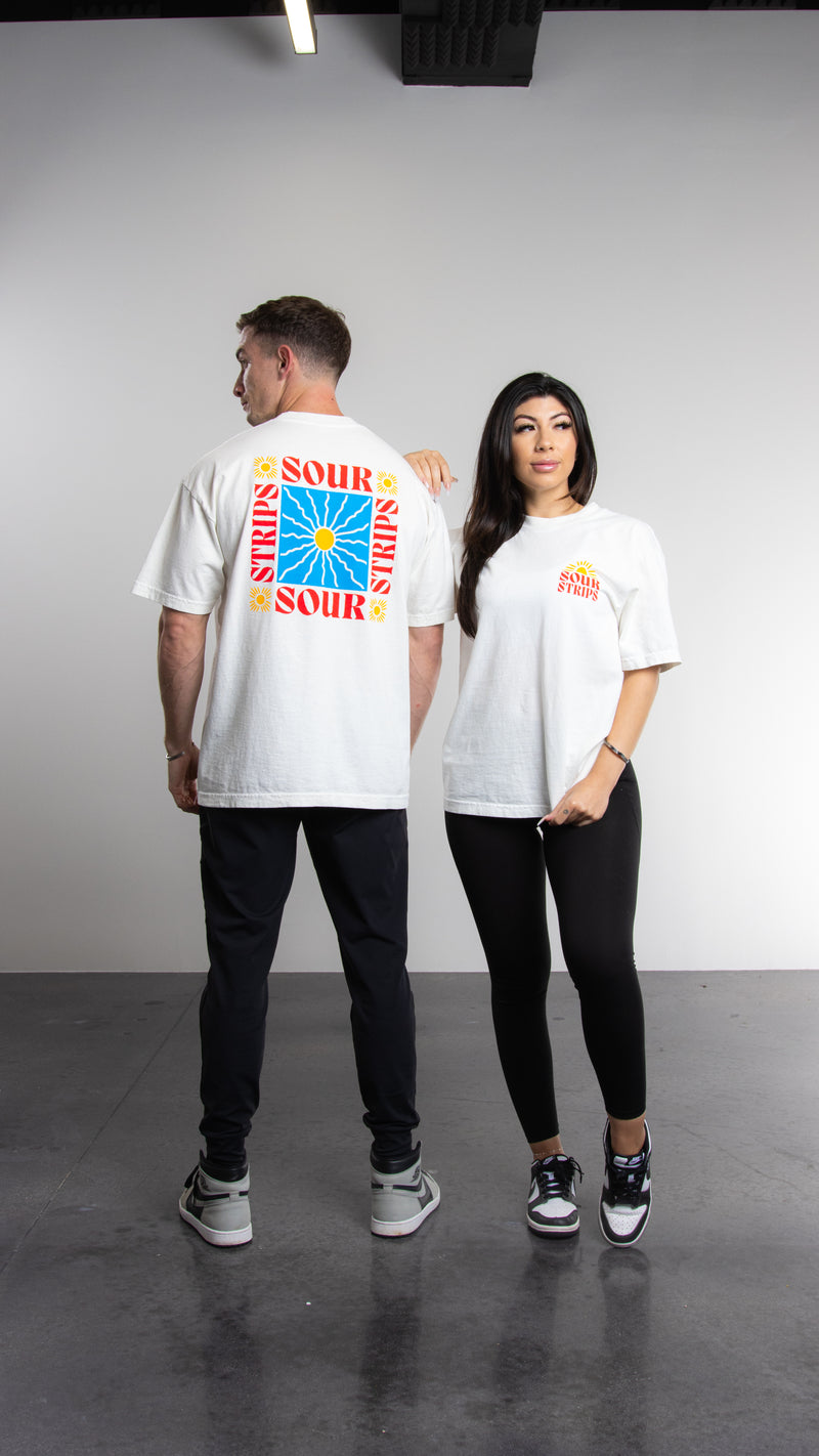 Two individuals pose in casual attire against a grey backdrop. The person on the left is facing away, revealing vibrant graphics and text saying "SOUR" on the back of their oversized box fit Sunshine Tee from SourStrips. The person on the right faces forward, wearing a white Sunshine Tee with subtle text on the front, showcasing a unisex design by SourStrips.