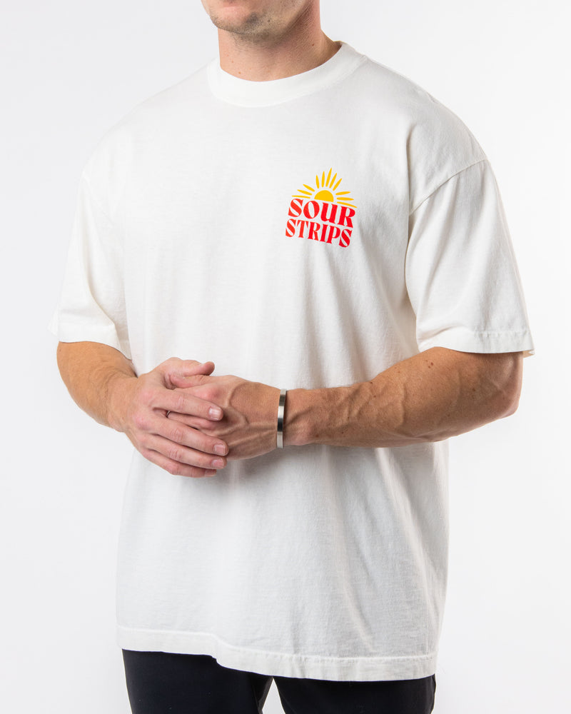 A person wearing the "Sunshine Tee" by SourStrips, which features a sun graphic on the chest and "Sour Strips" text, stands against a plain white background. The unisex design boasts an oversized box fit, with the person's hands loosely clasped in front while their face remains out of frame.