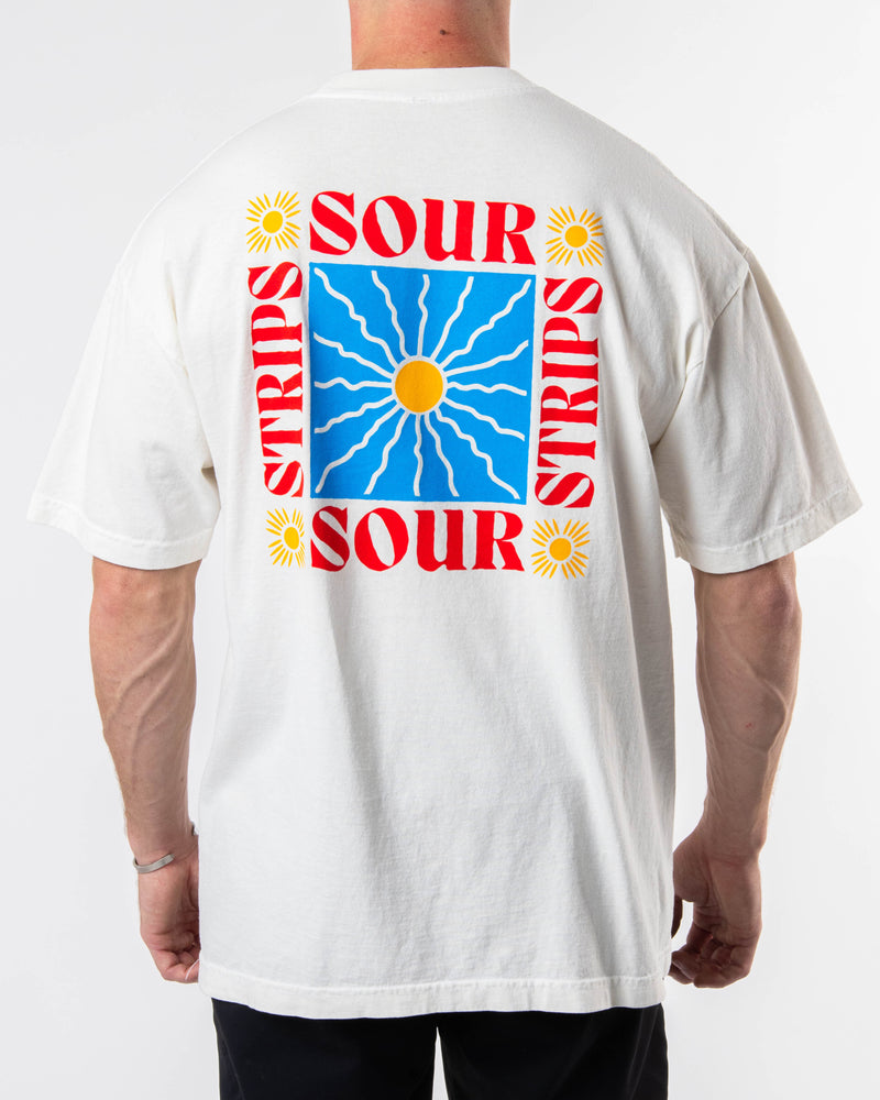 A person with short hair is shown from the back wearing a white oversized box fit Sunshine Tee by SourStrips. This unisex design features a colorful graphic on the back, showcasing a sun in a blue square with wavy lines, surrounded by the words "SOUR STRIPS" in red and yellow flower-like designs.