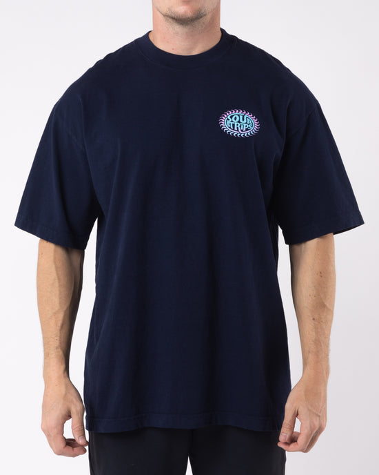 The individual is sporting the SourStrips Solar Tee, a dark blue oversized box fit T-shirt featuring a small, circular pink and teal graphic on the left chest area. The unisex design shows their hands relaxed by their sides, with only the upper body visible against a plain white background.