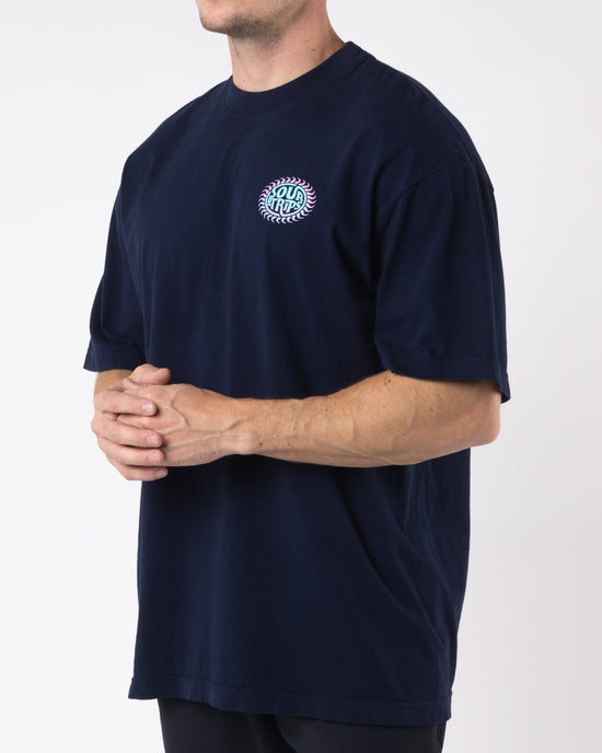 A person is standing against a plain background, wearing an oversized box fit Solar Tee in navy blue from SourStrips, featuring a colorful circular logo on the left side of the chest. The unisex design ensures versatility. They hold their hands clasped in front of them, with their face not visible in the image.