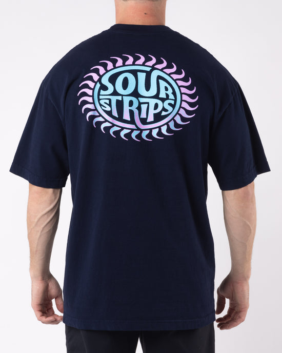 A person stands facing away, wearing an oversized box fit black Solar Tee from SourStrips. The unisex design features a colorful graphic on the back with the text "SOUR STRIPS" in blue and pink within a circular design bordered by a pattern of pink crescents. The background is plain white.
